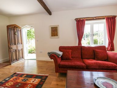 A living room with a red sofa red curtains a patterned rug wooden floor a wooden coffee table a framed picture on the wall and an open wooden door at Caldhu Cottage Holmrook