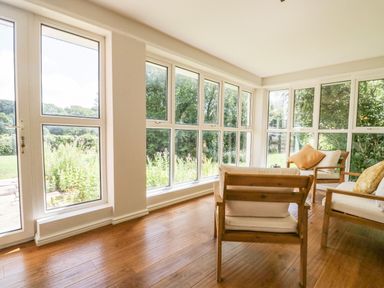 A sunroom with wooden chairs with cushions and large windows showing a garden at Caldhu Cottage in Holmrook