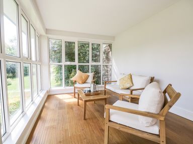 A sunroom with wooden chairs and a sofa with white cushions and pillows a wooden coffee table and large windows showing greenery outside at Caldhu Cottage in Holmrook