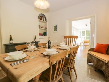 A dining room with a wooden table set with bowls plates glasses and cutlery wooden chairs a wicker bench with a cushion and a view into a sunroom at Caldhu Cottage in Holmrook