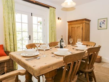 A dining room with a wooden table set with bowls plates cutlery and wine glasses wooden chairs and a cupboard with green curtains on windows at Caldhu Cottage in Holmrook