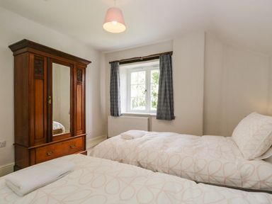 A bedroom with two single beds a wooden wardrobe and a window with curtains at Caldhu Cottage in Holmrook