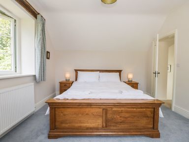 A bedroom with a wooden bed frame, white bedding, two bedside tables with lamps, a window with curtains, and an open door at Caldhu Cottage in Holmrook