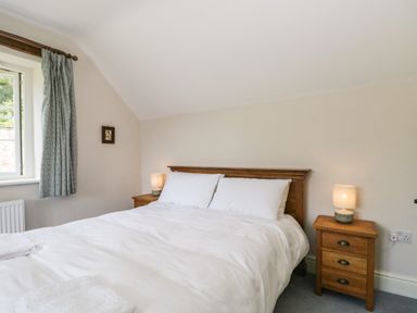 A bedroom with a double bed with white bedding two wooden bedside tables with lamps and a window with patterned curtains at Caldhu Cottage in Holmrook