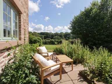 A garden patio with two wooden chairs with white cushions and a wooden table by a brick wall at Caldhu Cottage in Holmrook