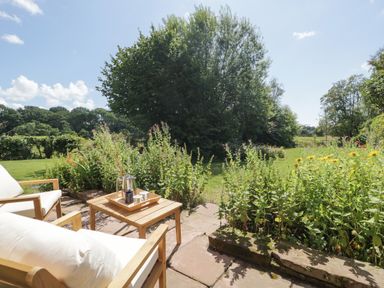 A garden patio with wooden chairs and a table holding a coffee pot and cups surrounded by flowers and grass at Caldhu Cottage in Holmrook