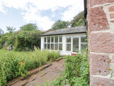 A garden with flowers and greenery next to a white conservatory attached to a red brick building at Caldhu Cottage in Holmrook