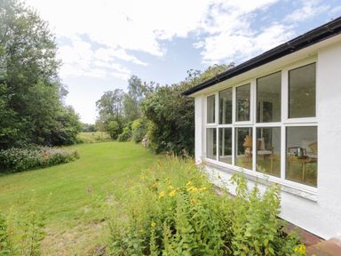 A garden with grass and flowers next to a white building with large windows at Caldhu Cottage in Holmrook