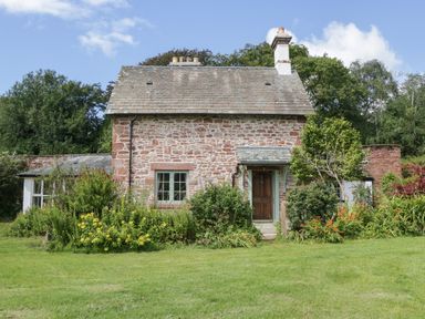 A stone cottage with a tiled roof surrounded by shrubs and flowers with a lawn in front at Caldhu Cottage in Holmrook