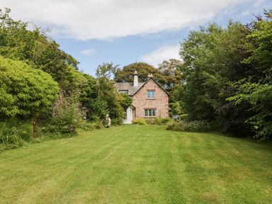 A brick house with two chimneys surrounded by green trees and grass at Caldhu Cottage in Holmrook