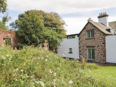 A stone and white cottage with green window frames surrounded by greenery and a garden at Caldhu Cottage in Holmrook