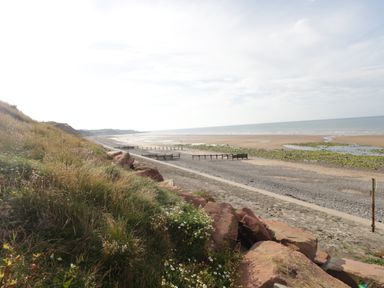 A beach with sand and pebbles next to grassy cliffs and a cloudy sky at Caldhu Cottage in Holmrook
