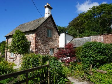 A stone cottage with a slate roof surrounded by green bushes and trees at Caldhu Cottage in Holmrook
