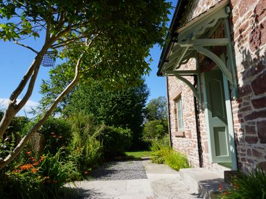 The exterior of a stone house with a green door canopy and surrounding garden with trees and flowers at Caldhu Cottage in Holmrook