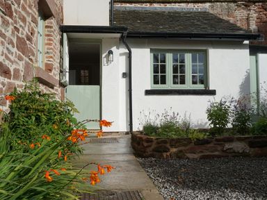 A garden path leading to a half open green door next to a white wall with green windows and flower beds at Caldhu Cottage in Holmrook