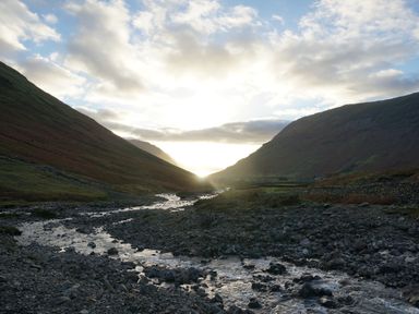 A rocky river flowing between two hills under a cloudy sky with the sun shining through the valley at Caldhu Cottage in Holmrook