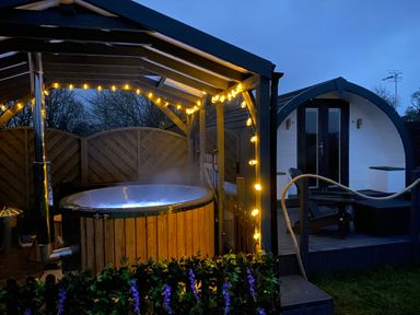 An outdoor covered hot tub with string lights and a wooden pod cabin in the background at Cherry Pod in Hookagate near Meole Brace