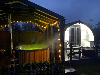 An outdoor area with a hot tub under a roof with string lights and a small pod with double doors and outdoor seating at Cherry Pod in Hookagate near Meole Brace