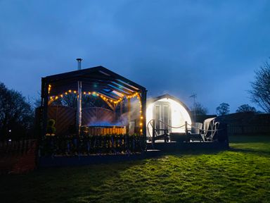 An outdoor area with a hot tub under a covered structure with string lights and chairs near a pod at Cherry Pod in Hookagate near Meole Brace