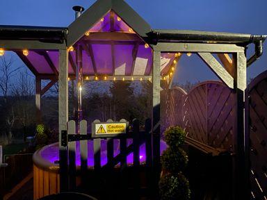 An outdoor wooden hot tub with purple lighting under a wooden gazebo with string lights at Cherry Pod in Hookagate near Meole Brace