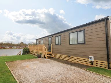 A brown mobile home with windows and a wooden deck near a lake with grass and gravel at Elim South Lakeland Leisure Village near Tewitfield