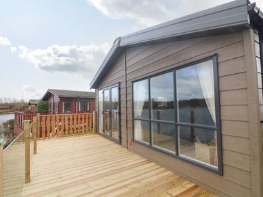 A wooden deck with glass railing attached to a cabin overlooking a body of water at Elim South Lakeland Leisure Village near Tewitfield