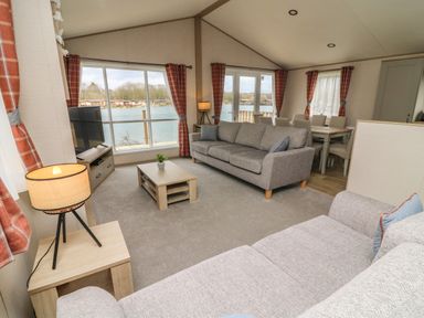 A living room with sofas a coffee table a TV and dining table near large windows overlooking a lake at Elim South Lakeland Leisure Village near Tewitfield