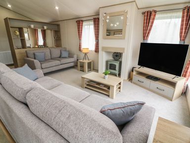 A living room with two sofas a coffee table a television on a stand a small fireplace and windows with curtains at Elim South Lakeland Leisure Village near Tewitfield