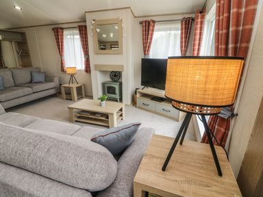 A living room with grey sofas a coffee table a TV on a stand a tripod lamp and red plaid curtains at Elim South Lakeland Leisure Village near Tewitfield