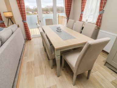 A dining area with a rectangular wooden table and eight upholstered chairs near glass doors overlooking water at Elim South Lakeland Leisure Village near Tewitfield