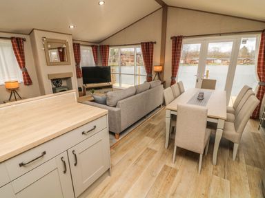 An open living and dining area with a dining table and chairs a sofa a television and large windows overlooking water at Elim South Lakeland Leisure Village near Tewitfield