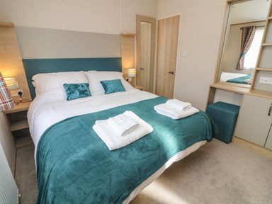 A bedroom with a double bed folded towels teal bedspread and pillows bedside tables lamps a mirror and a window with plaid curtains at Elim South Lakeland Leisure Village near Tewitfield