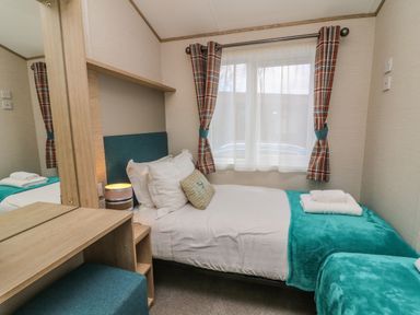 A bedroom with a single bed with white pillows and teal blanket near a window with curtains and a dressing table with stool at Elim South Lakeland Leisure Village near Tewitfield