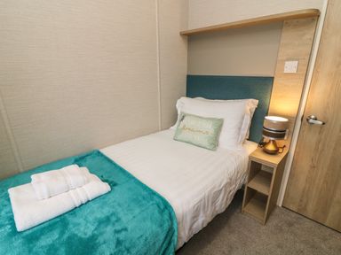 A small bedroom with a single bed covered with white sheets and a teal blanket with folded towels on top next to a wooden nightstand with a lamp at Elim South Lakeland Leisure Village near Tewitfield