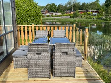An outdoor deck with a dining table set with chairs and a bottle with glasses overlooking a pond at Elim South Lakeland Leisure Village near Tewitfield