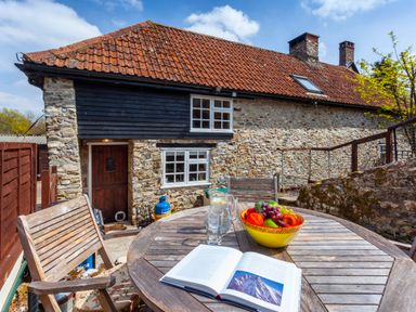 A round wooden table with a fruit bowl open book and glasses on a patio outside a stone cottage at Southcott Farm Cottage in Sheldon