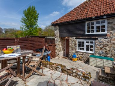 An outdoor patio with wooden table and chairs a barbecue grill and a stone wall next to a stone and wood house at Southcott Farm Cottage in Sheldon
