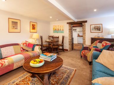 A living room with patchwork sofas a wooden coffee table with fruit and books and a dining table with chairs at Southcott Farm Cottage in Sheldon