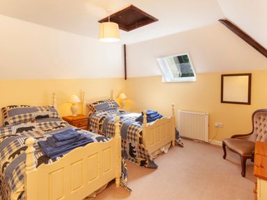 A bedroom with two single beds blue patterned bedding wooden nightstand with lamps radiator window and a chair at Southcott Farm Cottage in Sheldon