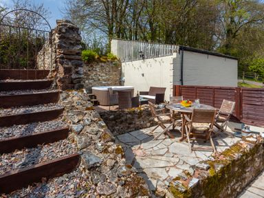 An outdoor patio area with wooden chairs around a round table and a hot tub with seating near a stone wall at Southcott Farm Cottage in Sheldon