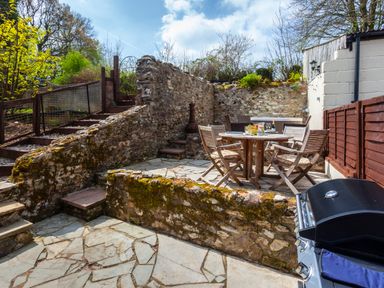 An outdoor patio with stone walls wooden table and chairs and a barbecue at Southcott Farm Cottage in Sheldon