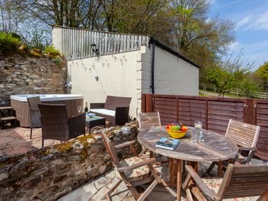 An outdoor patio area with wooden round table and chairs a hot tub and garden chairs at Southcott Farm Cottage in Sheldon
