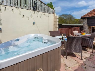 An outdoor patio with a hot tub a table with chairs and a bottle of champagne at Southcott Farm Cottage in Sheldon