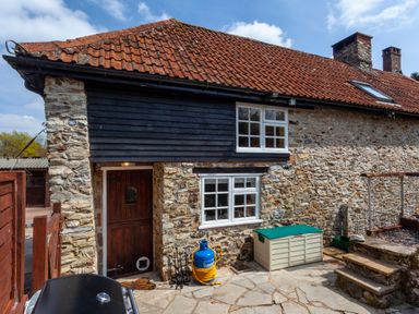 Stone cottage with a tiled roof patio area wooden door two white framed windows gas cylinder and storage box at Southcott Farm Cottage in Sheldon