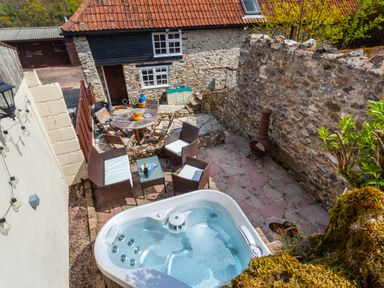 A patio with a hot tub seating area and table set next to a stone wall at Southcott Farm Cottage in Sheldon