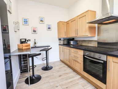 A kitchen with wooden cabinets a built in oven a black countertop a small dining area with two black stools and framed pictures on the wall at Aryelle House in Kendal