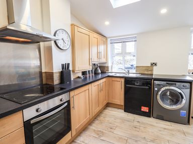 A kitchen with wooden cabinets a black countertop a built in oven stove dishwasher washing machine and a wall clock at Aryelle House in Kendal