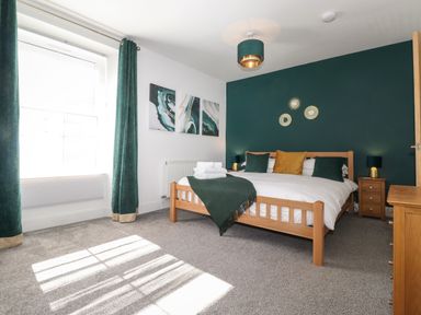 A bedroom with a wooden bed with white bedding green and mustard cushions green curtains and three wall art pieces at Aryelle House in Kendal