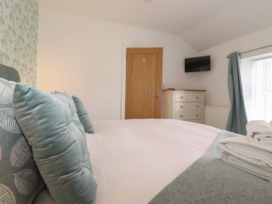 A bedroom with a bed with blue pillows folded towels on the bed a wooden door a chest of drawers a wall-mounted TV and a window with curtains at Aryelle House in Kendal