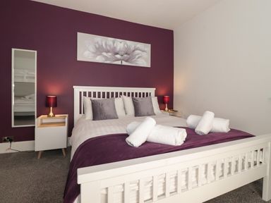 A bedroom with a white bed frame purple accent wall bedside tables with lamps and rolled towels on the bed at Aryelle House Kendal